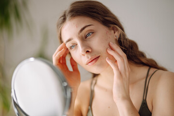 Beautiful woman looking in the mirror, touching her face with her hand, examines acne,...