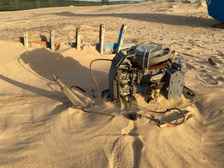 A rusty dismantled boat engine lies partially in the sand on the beach. The wooden structure of an abandoned wrecked boat is bathed in warm sunlight.