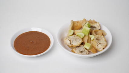 A portion of batagor (fried tofu meatballs) topped with sliced ​​cucumber served in a white container with peanut sauce in a separate container placed on a white base and background (isolated white).