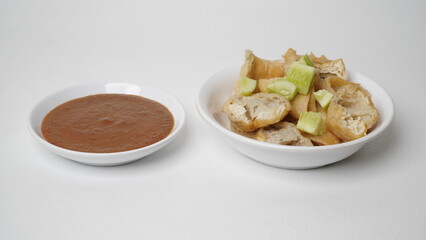 A portion of batagor (fried tofu meatballs) topped with sliced ​​cucumber served in a white container with peanut sauce in a separate container placed on a white base and background (isolated white).