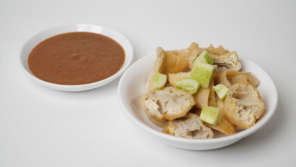 A portion of batagor (fried tofu meatballs) topped with sliced ​​cucumber served in a white container with peanut sauce in a separate container placed on a white base and background (isolated white).