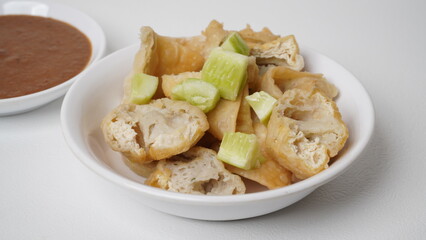 A portion of batagor (fried tofu meatballs) topped with sliced ​​cucumber served in a white container with peanut sauce in a separate container placed on a white base and background (isolated white).