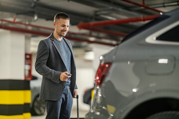 A smart casual businessman is standing at garage with luggage and opening car trunk with key while...
