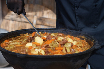 Cooking thick meat soup in a large cauldron outdoors. Catering concept. Chef stirs rich meat broth with vegetables. Close-up. Onion and carrot. Orange bright saturated color.