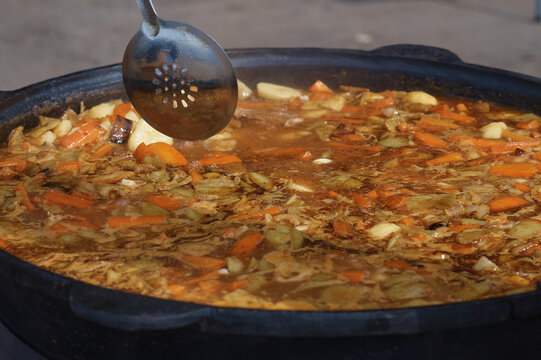 Cooking thick meat soup in a large cauldron outdoors. Catering concept. Chef stirs rich meat broth with vegetables. Close-up. Onion and carrot. Orange bright saturated color.