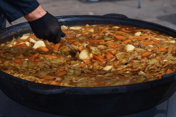 Cooking thick meat soup in a large cauldron outdoors. Catering concept. Chef stirs rich meat broth with vegetables. Close-up. Onion and carrot. Orange bright saturated color.