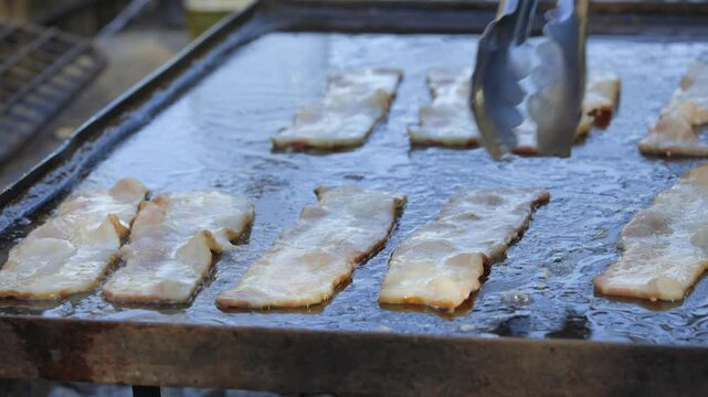 Tongs flip fatty bacon strips cooking, sizzling on hot outdoor griddle