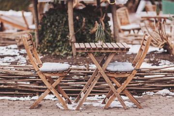 A cozy wooden table and chairs sit in a winter wonderland, lightly dusted with snow. A small...