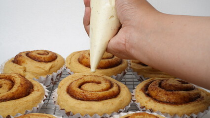 A hand is topping cream cheese frosting on a collection of cinnamon rolls using a paper cup placed on a cooling rack and against a white background (isolated white).