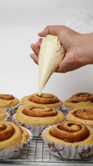 A hand is topping cream cheese frosting on a collection of cinnamon rolls using a paper cup placed on a cooling rack and against a white background (isolated white).