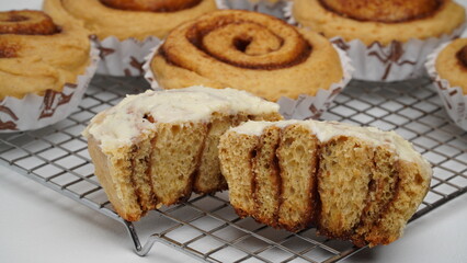 A collection of cinnamon rolls using paper cups and one of them is topped with cream cheese frosting and placed on a cooling rack with a white background (isolated white).