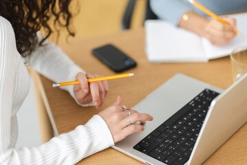 A close-up of a person wearing a white sweater using a laptop and holding a pencil, with a smartphone and notebook on a wooden desk in a bright workspace.