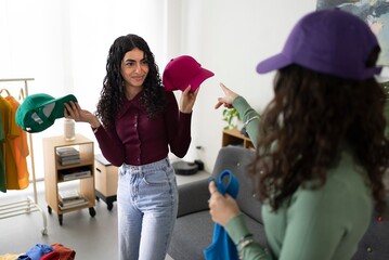 Two young women with curly hair are engaged in a discussion about hat colors, one holding a green and magenta cap while the other points, standing in a stylish boutique setting.