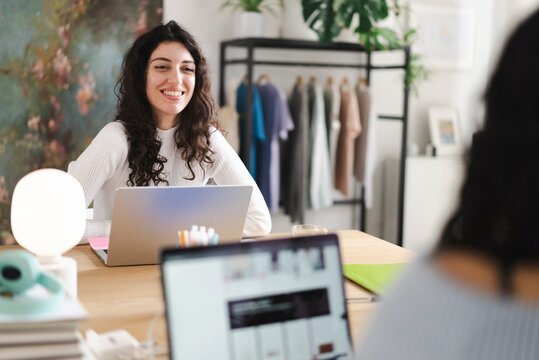 A young woman with curly hair wearing a white sweater is smiling while working on her laptop in a stylish workspace with clothing racks and decorations in the background.
