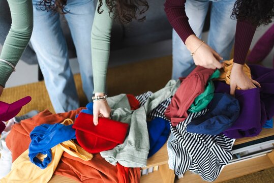 Two young women are searching through a pile of colorful clothing items on a wooden table, selecting pieces in a stylish and trendy boutique setting.
