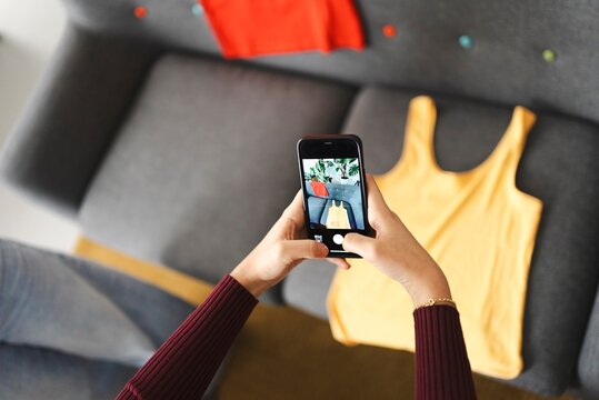 A woman in a burgundy sweater is holding a smartphone and capturing a photo of a yellow top placed on a couch, surrounded by a stylish and trendy setting.