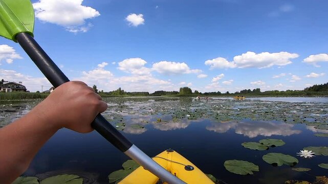 Kayaking along river through growing white lily nymph on sunny summer day. Person floating in boat on river with surface of water reflecting white clouds on calm day. Tourism. Active lifestyle. POV