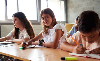 Fototapeta premium Portrait of a cheerful teenage girl looking at camera smiling while taking a test in elementary school sitting next to colleagues. Image with copy space.