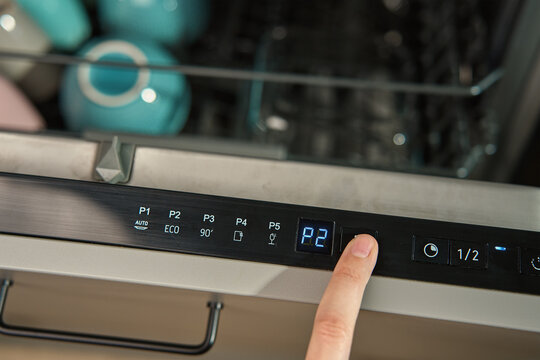 Close-up of female finger pressing button on dishwasher control panel. Woman using dishwasher in modern kitchen. Modern kitchen appliance with digital settings for efficient dishwashing