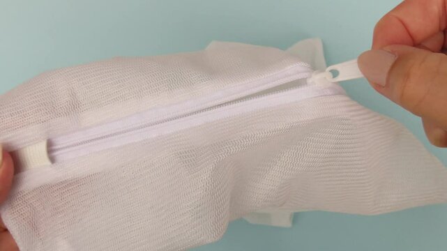 Closeup video of woman hands showing a white zipped laundry bag, how to open and closing it, and its material. Blue background. Mesh laundry bag.