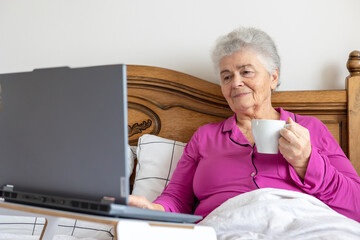 Fototapeta premium Senior woman relaxing in bed with laptop and tea, enjoying morning comfort and digital connection. Copyspace