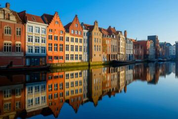 Obraz premium Traditional colorful houses reflecting on river in Belgium, creating a picturesque cityscape under the clear blue sky of a sunny morning