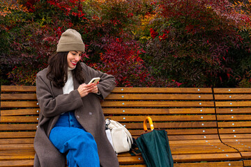 A relaxed young woman sitting on a bench in the park, with autumn-colored plants behind her, smiling while scrolling through her mobile phone. Copy space
