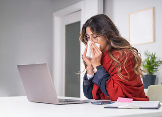 Businesswoman blowing nose, side view portrait of caucasian sick businesswoman blowing nose in office sitting desk wrapped scarf freezing. Sinus allergies concept idea image, copy space.
