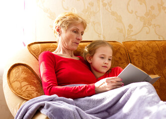 Grandmother and granddaughter reading a book sitting on the couch and covered with a plaid. A cozy...