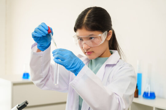 Caucasian school children learning experiment in science classroom. Young female scientist in protective goggles analyzing a microscope sample while holding a tablet for research documentation.
