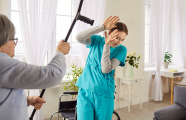 Aggressive elderly woman threatening with crutch to frightened nurse have disagreement being...