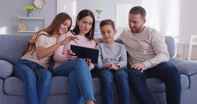 Happy young family with two little kids at home, relaxing on sofa in living room using tablet online, talking speak on video webcam call on computer, parents with teen children at using gadget