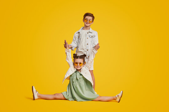 Trendy little girl doing splits while posing happily with boy standing behind and having fun together on vivid yellow background