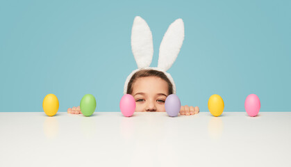Cute little girl with white bunny ears looking at camera while peeking out from behind table with colorful Easter eggs on blue background