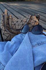 Sunglasses, denim jacket and women's handbag on wooden surface outdoors. Women's accessories