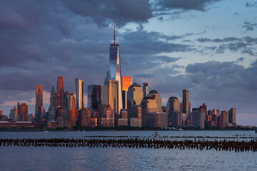 New York City skyline at sunset with Lower Manhattan and World Trade Center skyscrapers illuminated by golden light. Urban landscape across the Hudson River with Financial District and Battery Park