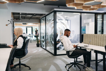Fototapeta premium In a modern office setting, an African American businessman and his Muslim colleague, wearing a hijab, engage in collaborative discussions, tackling various business tasks and solving problems