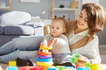 Caring happy mother or young babysitter playing pyramid toy with little daughter sitting on warm floor at home, family spending leisure time together, loving mum with baby kid having fun together