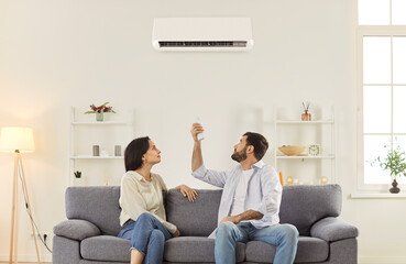 Young family couple sitting together on sofa in living room and turning on air conditioner. Focused...