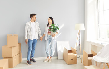Happy young smiling couple standing with cardboard boxes in new apartment holding hands and looking at each other celebrating moving day. Relocating, real estate, mortgage concept.