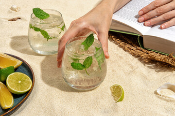 Two cold and refreshing cocktail with ice garnished mint standing on the sand, on the beach by the sea. Summer mint non-alcoholic cocktail with juice and soda. Hard light, bar menu.