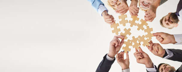 Bottom view of confident business people putting together pieces of wooden puzzles. Colleagues join jigsaw pieces isolated on white background. Cooperation, teamwork, help and support concept. Banner