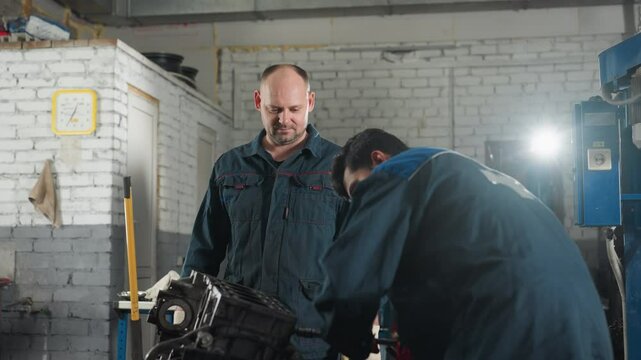 Professionals in mechanical workshop working on car engine, tightening nut with red gloves while colleague observes, background features work tools, yellow wall clock, and bright ligh