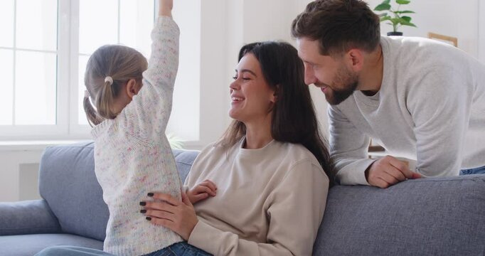 Cheerful excited parents listening to beloved kid girl, playing with child on sofa in modern home interior, together laughing, enjoying family leisure, baby talking, telling funny story to parents 