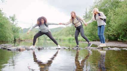 Multi-Cultural Group Of Female Friends Crossing River On Stepping Stones Hiking In UK Lake District