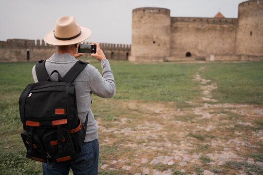 Senior male traveler in hat taking picture of old fortress on smartphone during vacation.