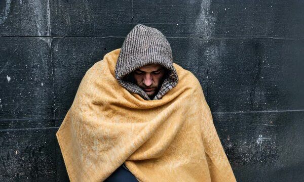 A somber image of a homeless individual sitting against a wall, wrapped in a blanket, highlighting the stark reality of urban poverty and the need for compassion in society.