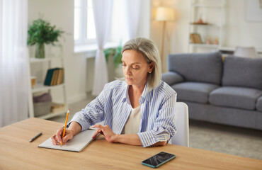 Retired old senior woman, pleasant elderly person alone at home, female sitting writing at desk,...