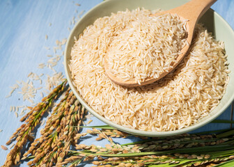 A bowl of brown-rice is placed on a table next to a spoonful and ears of rice.Rice is spread out in the bowl and the spoon is full of raw-rice and sitting on top of it