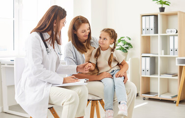 Friendly pediatrician with clipboard talking to little girl patient sitting on knees of her mother...